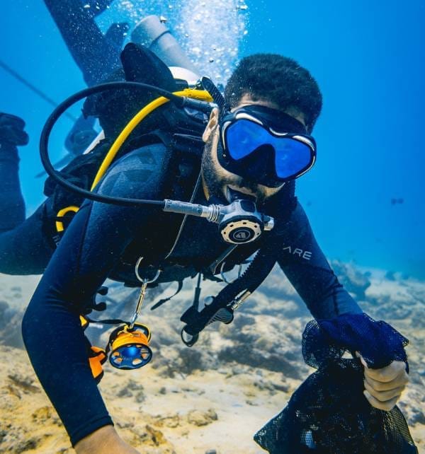 Scuba diver in Nusa Penida wearing blue mask and wetsuit collects debris into a mesh bag during an underwater cleanup.