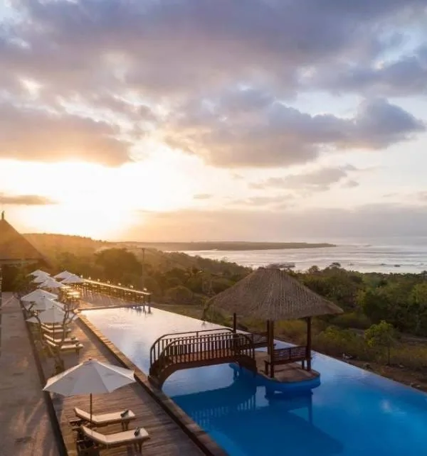 An Island Bursting with Adventures 1 Sunset lights an infinity pool at a Nusa Penida resort, sun reflecting on water beside a wooden bridge, thatched pool gazebo.