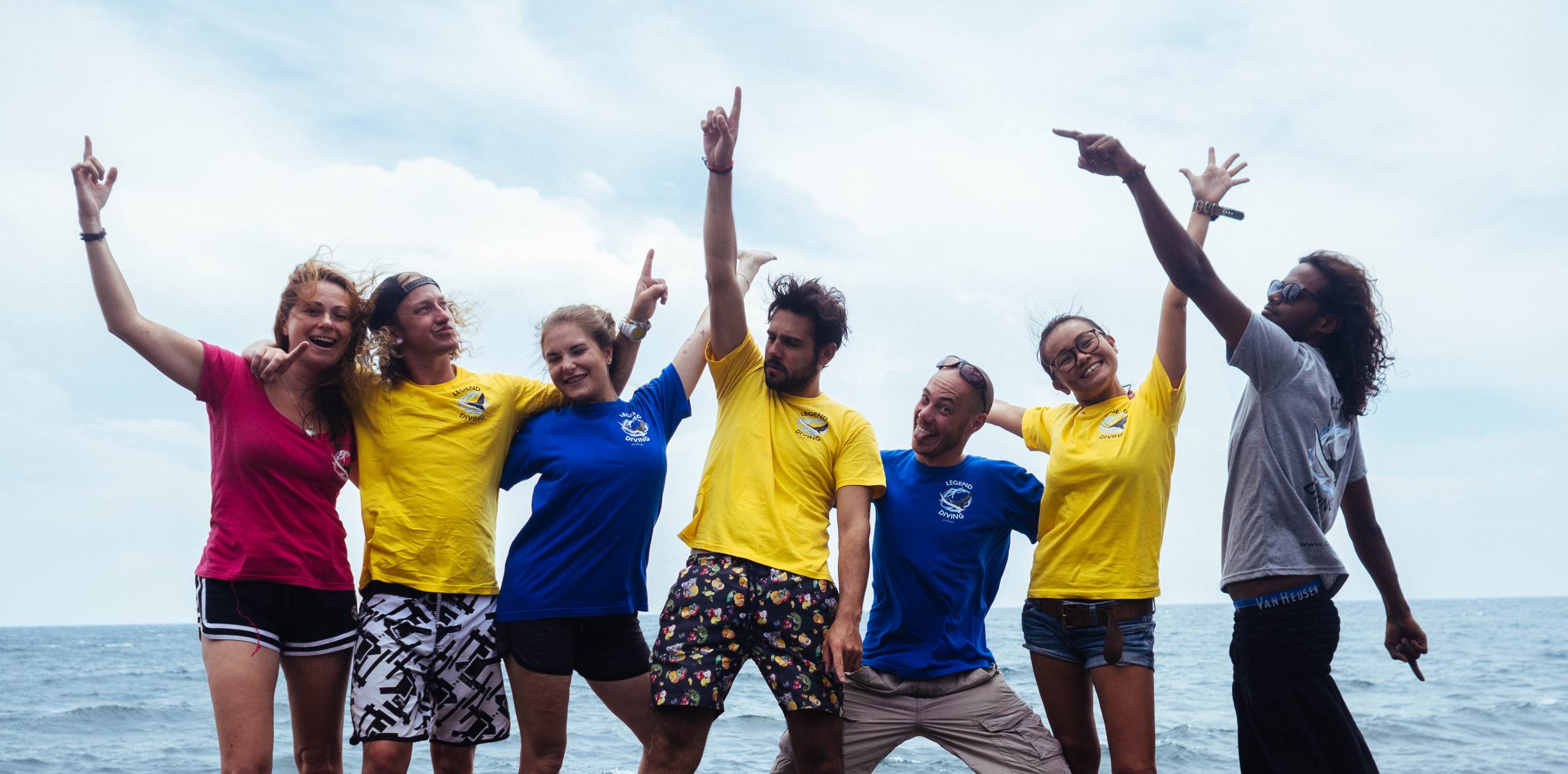 Divemaster Penida Header Grupo de siete personas en Nusa Penida celebrando en la orilla del mar, brazos alzados y sonrisas tras una inmersión.
