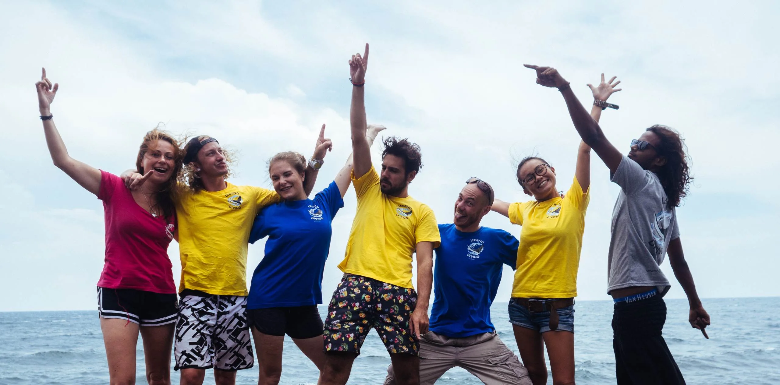 Divemaster Penida Header Seven people on Nusa Penida pose and cheer with arms raised, smiling against the ocean backdrop.