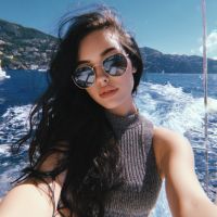Woman with long dark hair wearing round sunglasses and a sleeveless knit top taking a selfie on a boat with wake and coastal hills behind her.
