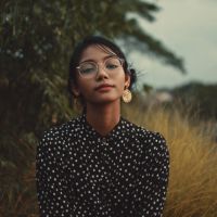 Young woman with round glasses and a single yellow flower earring wearing a black polka-dot blouse, seated outdoors among tall grass and trees.