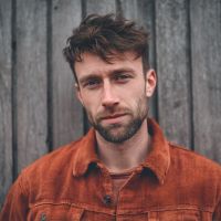 Young man with tousled brown hair and short beard wearing a rust-colored jacket, standing against a wooden plank wall.