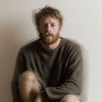 Bearded man with messy hair wearing an oversized dark sweater, seated hugging a knee against a neutral background, looking at the camera.