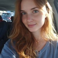 Red-haired woman with green eyes and light freckles smiling in a car with sunlight on her face.