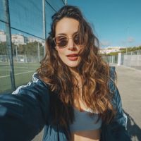 Young woman in sunglasses and a blue jacket takes a sunny selfie on a fenced sports court