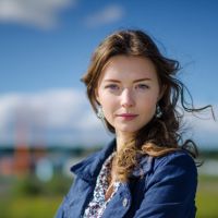 Young woman with wavy brown hair and earrings in a navy jacket and printed blouse, outdoors with hair blown by a breeze.