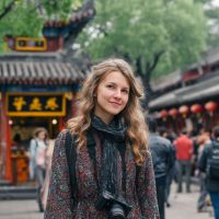 Young woman with wavy blonde hair and a camera around her neck, standing in a crowded Chinese street decorated with red lanterns.