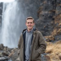 Smiling young man in a jacket and gray sweater stands on rocky ground with a tall waterfall and mossy cliff behind him.
