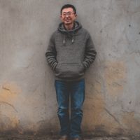 Man in a gray hoodie and jeans, hands in pockets and smiling while standing against a weathered concrete wall.