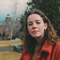 Woman with curly brown hair and hoop earrings wearing a red coat on a grassy campus with a brick building in the background.