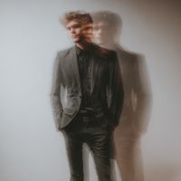 Man in dark suit with hands in pockets, shown in double-exposure blur that creates a ghosted second silhouette.