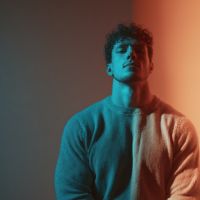 Man in a sweater lit by teal and orange split light, looking upward against a dark gradient background.