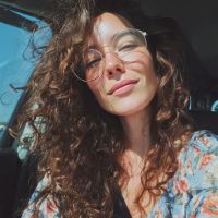 Sunlit selfie of a woman with curly hair and round glasses in a car, wearing a floral top