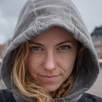 Close-up of a person with light brown hair wearing a gray hooded jacket, looking into the camera with a direct gaze.